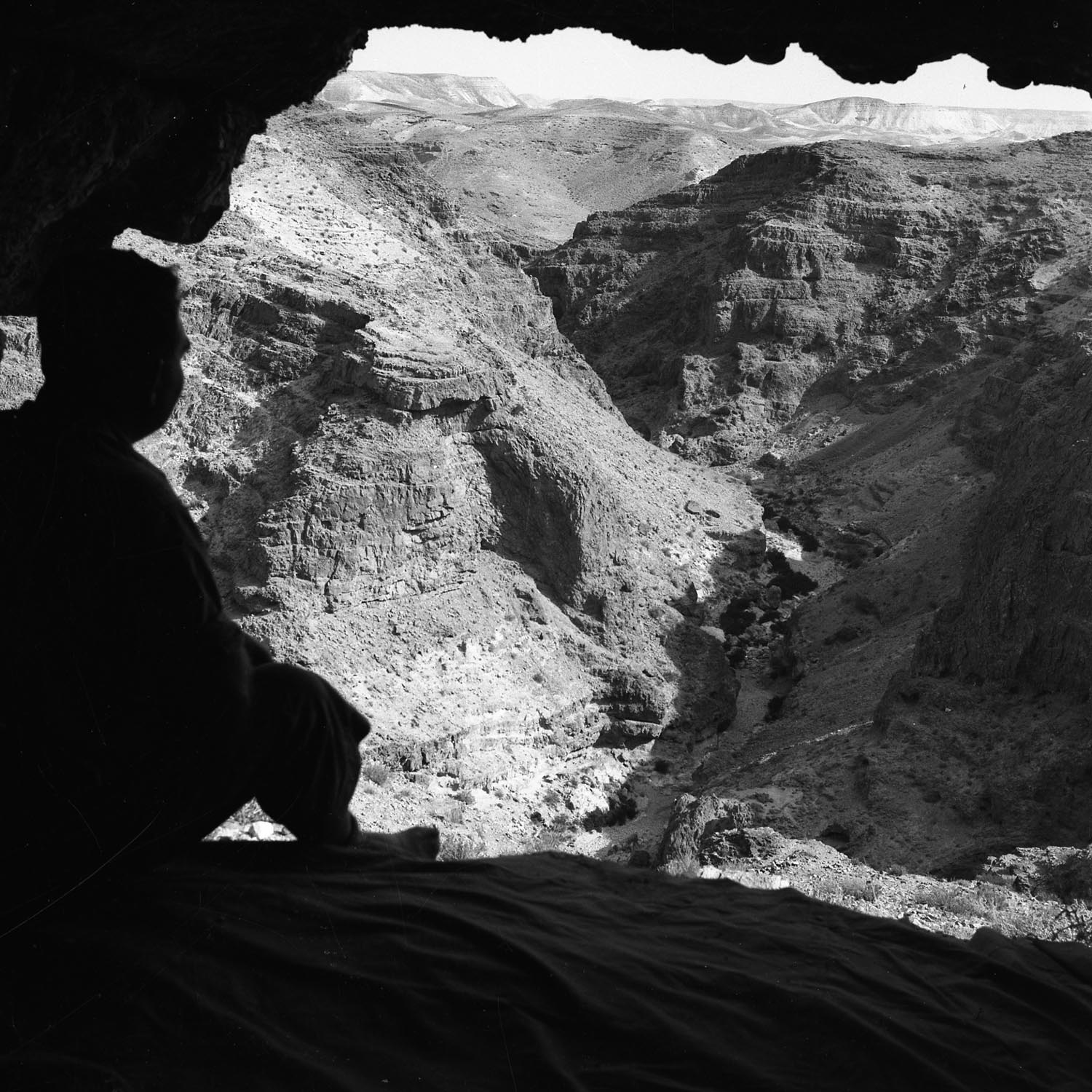 Foto em preto e branco tirada da boca de uma caverna, com a silhueta de uma pessoa e um vale desértico profundo ao fundo.